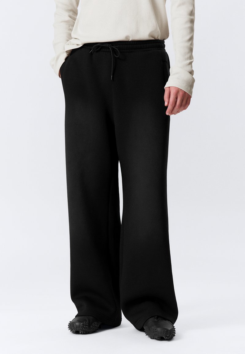 Μαύρο παντελόνι φόρμας με φαρδιά μ πατζάκια και ελαστική μέση με κορδόνι, από απαλό βαμβακερό ύφασμα, με πλαϊνές τσέπες, σε συνδυασμό με υφασμένα μαύρα παπούτσια.