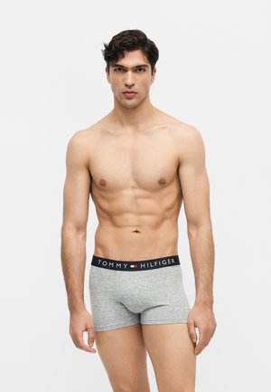 Młody mężczyzna bez koszulki z krótkimi ciemnymi włosami, w szarych bokserkach Tommy Hilfiger, stojący na jednolitym tle.