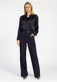 Zwarte, gestructureerde lange mouwen overhemd met knopen, gecombineerd met op maat gemaakte marineblauwe broek met een riem. Het shirt heeft een hoge kraag en een subtiele glans.