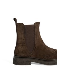 Bottine en suede marron avec des panneaux latéraux élastiques, des détails brogues et une semelle en caoutchouc texturée. Langue à l'arrière pour un enfilage facile.
