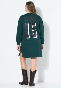 Vestido sudadera verde con lettering negro "STUDIO 15" y aberturas laterales. Combinado con botas de cowboy marrones hasta la rodilla. Textura de tela suave.
