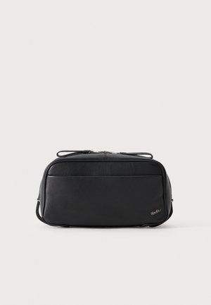 Trousse de toilette rectangulaire en cuir noir, avec une fermeture éclair sur le dessus et de petits éléments sur les côtés, sur un fond clair uni.