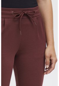 Vinröda sweatpants med dragsko i midjan, med en slät yta och sidofickor. Närbild av en hand som vilar på tyget.
