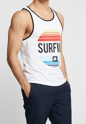 Homme portant un débardeur blanc sans manches avec des bordures noires, un texte coloré "SURFING", et un pantalon bleu foncé, les mains dans les poches.