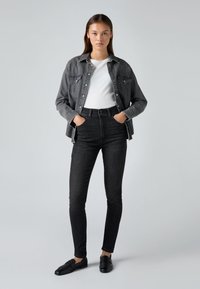 Harmaa denim-paita valkoisen t-paidan päällä, yhdistetty korkeavyötäröisiin mustiin farkkuihin ja mustiin loafereihin, esillä yksinkertaisen taustan edessä.