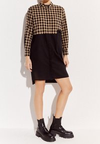 Czarna i brązowa, w kratkę sukienka koszulowa z kołnierzykiem zapinanym na guziki i długimi rękawami, z czarną dolną częścią i boczną kieszenią. Noszona z czarnymi botkami za kostkę.