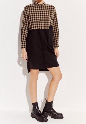 Czarna i brązowa, w kratkę sukienka koszulowa z kołnierzykiem zapinanym na guziki i długimi rękawami, z czarną dolną częścią i boczną kieszenią. Noszona z czarnymi botkami za kostkę.