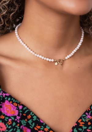 Perlenkette mit runden weißen Perlen, einem goldenen Verschluss und einem kleinen Charm, vor einem floralen Hintergrund.