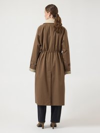 Ženska s čopom, ki nosi rjavi dolg plašč z izrazitim pasom, zavihane rokave, črne hlače in bež čevlje z visokimi petami, obrnjena stran.