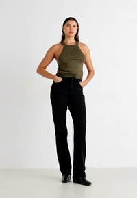 Olijfgroene haltertop met kleine versieringen, gecombineerd met hoge zwarte jeans. Model staat met handen in de zakken op een neutrale achtergrond.