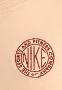 Peach fabric features a round, embroidered logo in brown reading "NIKE" and "THE SPORTS AND FITNESS COMPANY." Smooth, stretchy texture.