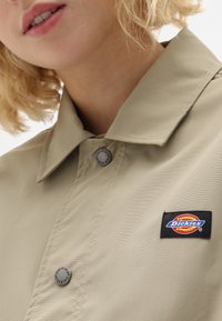 Beige knappjacka med spridd krage, med metallknappar och en rektangulär Dickies-logotyp på vänster sida. Slät textur.