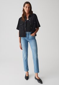 Cropped shirt nera con due tasche frontali, abbinata a jeans dritti azzurri chiari e scarpe slip-on nere. Outfit semplice e casual.