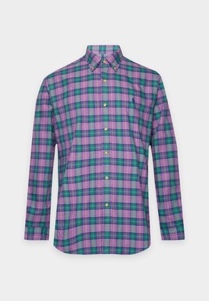 Un homme porte une chemise à carreaux violette, un pull bleu marine posé sur les épaules, un pantalon foncé, des chaussures noires et une casquette plate sur un fond uni.