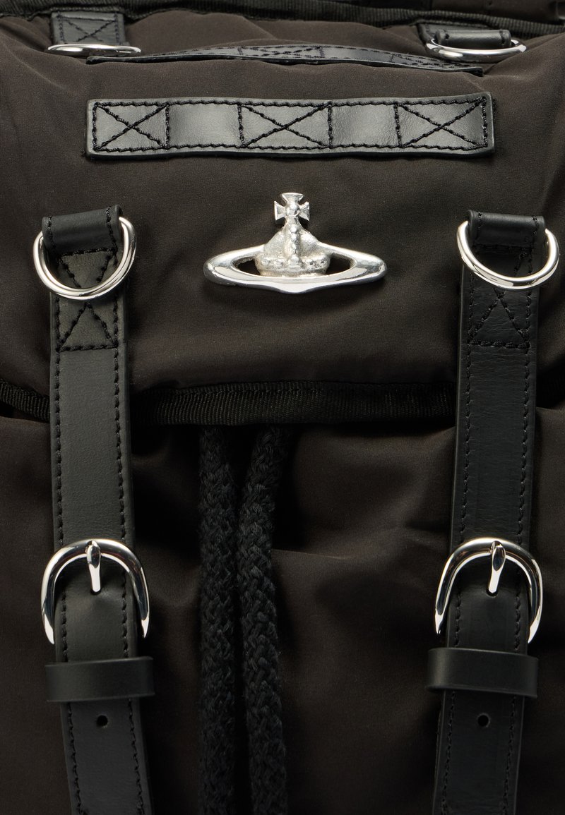 Sort rygsæk i stof med sølvfarvede metaldele, læderremme og en flettet snor til lukning. Inkluderer et karakteristisk metalemblem.