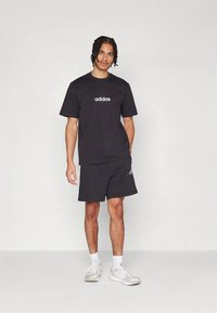 Μαύρο t-shirt Adidas με λευκό λογότυπο, συνδυασμένο με μαύρο σορτς που έχει πλευρικές ρίγες, φορεμένα με αθλητικά παπούτσια ανοιχτού χρώματος.