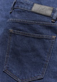 Tumedat sinised teksad, millel on must nahkpaber koos tõstetud logoga. Kontrastne õmblus tasku ja vöökoha lähedal annab detaili.