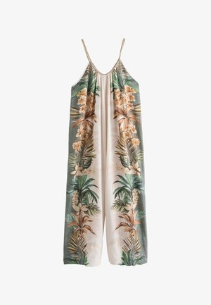 Joven mujer de pie sobre tierra arenosa, vistiendo un vestido largo y suelto con patrones florales tropicales, con palmeras al fondo.