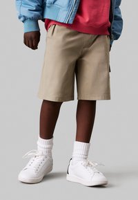 Pantaloni corti beige con cintura elastica, sneakers bianche con dettagli neri e calze bianche a coste; indossati con una giacca bomber blu chiaro.