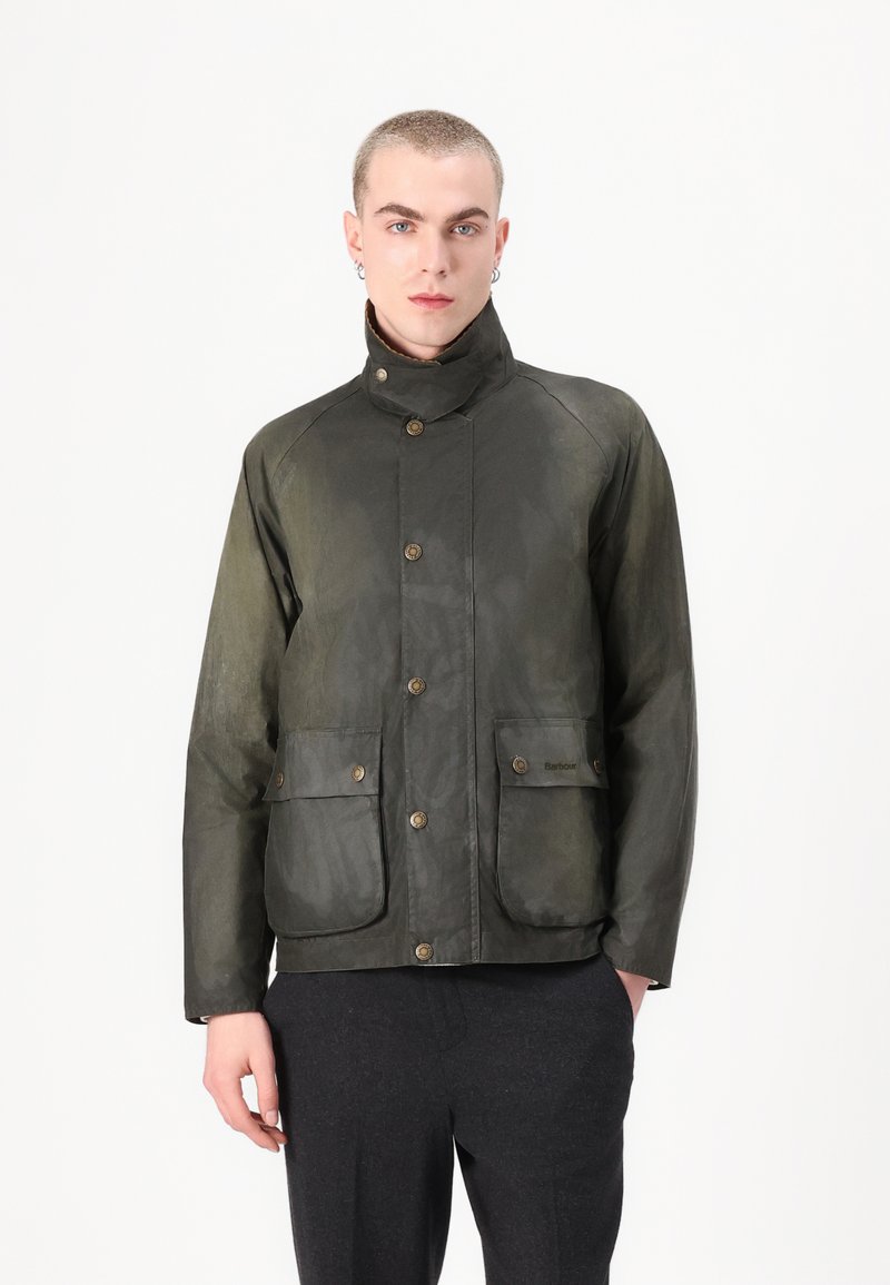 Casaco encerado verde-oliva com dois bolsos frontais, botões de latão, gola alta e uma superfície texturizada. Design slim e moderno.
