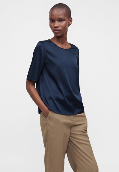 Femme aux cheveux courts portant un chemisier en satin bleu marine à manches courtes et un pantalon beige, les mains dans les poches, debout devant un fond uni.