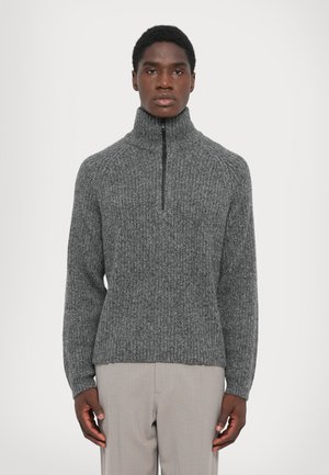 Man wearing a grey ribbed knit sweater with a high zip collar and light beige trousers, standing against a plain background.
