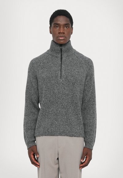 Man in een grijze geribbelde gebreide trui met een hoge ritskraag en lichtbeige broek, staande tegen een effen achtergrond.