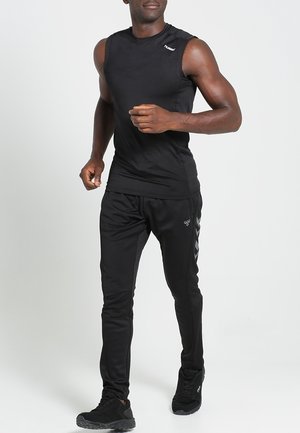Homme athlétique courant en débardeur noir, pantalon de survêtement noir avec un motif chevron sur le côté, et chaussures de course noires sur un fond uni.