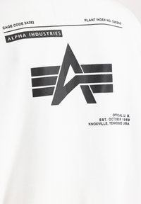 Bijela pamučna majica s crnim grafičkim printom koji sadrži slovo "A" i horizontalne pruge, te tekst koji detaljno opisuje brend i lokaciju.