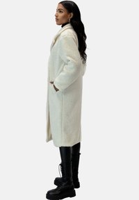 Lange, crème-kleurige jas van getextureerd, pluche materiaal, voorzien van zijzakken, een kraag en een rechte snit, gedragen over zwarte legging en laarzen.