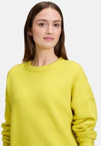 Żółty sweter z wełny z okrągłym dekoltem, fakturowaną tkaniną, opuszczonymi ramionami i dopasowanymi rękawami, prezentowany na neutralnym tle.