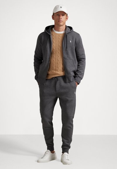 Sivi zip-up dukserica, bež pleteni pulover, sivi trenirci s uzicom i bijele tenisice. Sadrži logotip kao dekoraciju na odjeći.
