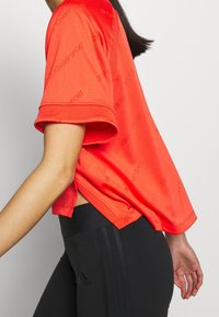 Czerwona sportowa koszulka z krótkim rękawem, ozdobiona powtarzającym się wzorem z logo "TOMMYSPORT". Boczne rozcięcia dla ułatwienia ruchów.