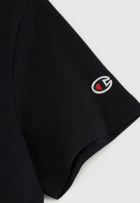 T-shirt in cotone nero a maniche corte, con un logo ricamato distintivo rosso e bianco sulla manica sinistra.