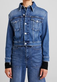 Skórzana kurtka w kolorze średniego niebieskiego z klasycznym kołnierzykiem, dwoma kieszeniami na piersi, srebrnymi zatrzaskami i kontrastowymi czarnymi mankietami.