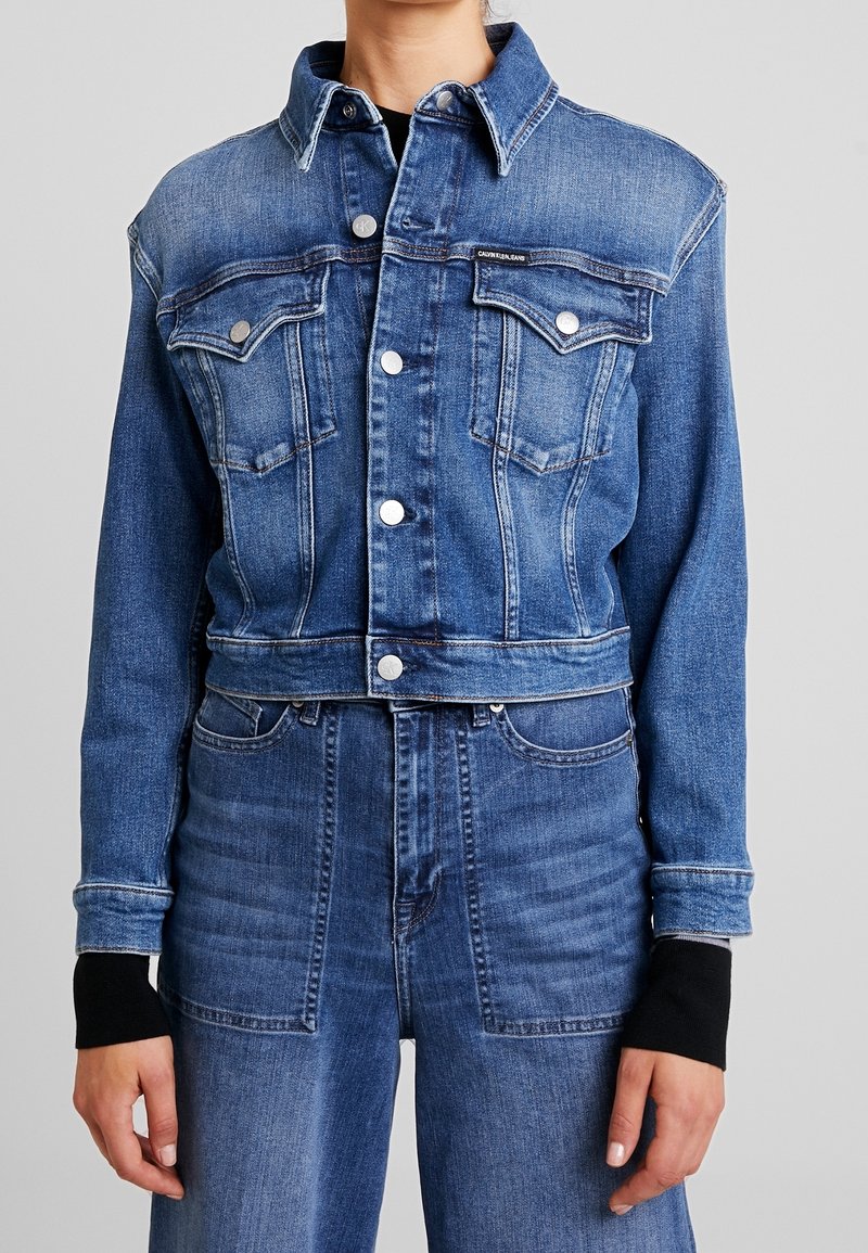 Skórzana kurtka w kolorze średniego niebieskiego z klasycznym kołnierzykiem, dwoma kieszeniami na piersi, srebrnymi zatrzaskami i kontrastowymi czarnymi mankietami.