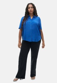 Blue short-sleeve shirt with button closure and a chest pocket, paired with black flared pants and sandals with a slight heel.