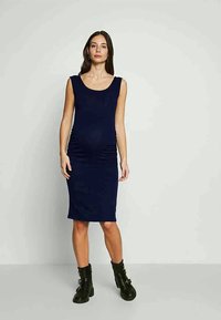 Mulher grávida em pé, vestindo um vestido azul-marinho sem mangas até ao joelho, com botas de tornozelo pretas com atacadores, contra um fundo simples.