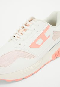 White sneaker with mesh and synthetic upper, featuring light pink accents, laces, and textured rubber outsole. Prominent branding detail on side.