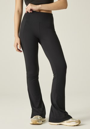 Femme portant un pantalon de yoga évasé noir et des chaussures de sport beiges, debout devant un fond clair.