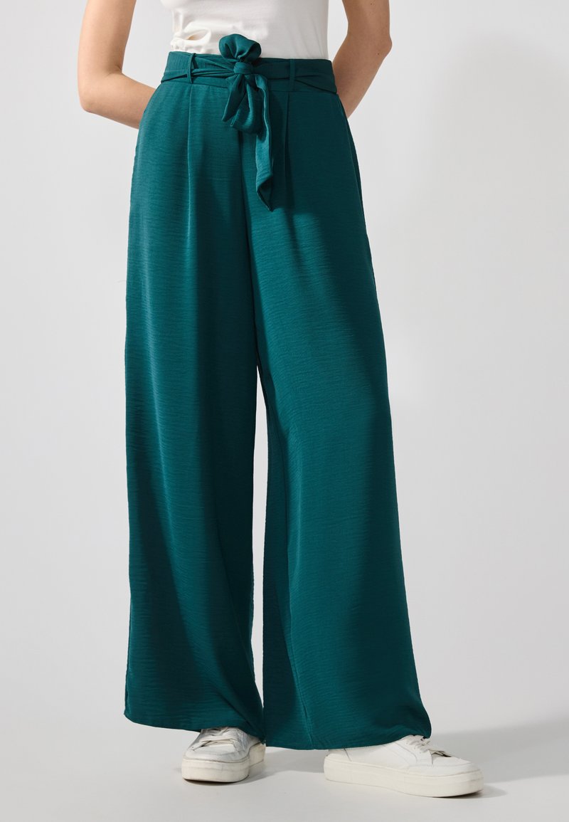 Pantalons larges teal en tissu texturé, dotés d'une ceinture nouée à la taille et d'une coupe décontractée, associés à des baskets blanches.