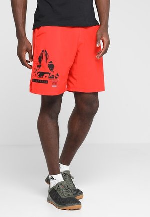 Homme portant un short de sport rouge vif avec un logo géométrique noir et des chaussures de sport gris foncé avec des chaussettes blanches Adidas, debout sur un sol gris.
