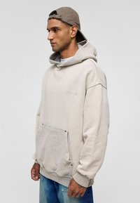 Beige hoodie met een trekkoord en een textuurzak. Materiaal lijkt zacht. Gecombineerd met vervaagde blauwe denim. Weergegeven op een neutrale achtergrond.