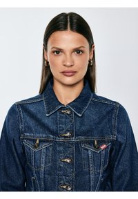 Otse tumedate sirgete juustega naine, kes kannab kuldseid rõngaskõrvarõngaid ja sinist teksajopet nuppude ja rinnataskutega, vaadates otse ette.