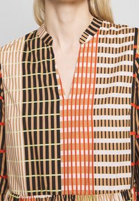 Bunt gemustertes Kleid mit einem geometrischen Rasterdesign in Braun, Orange, Weiß und Grün, V-Ausschnitt und gerafftem Taillendetail.