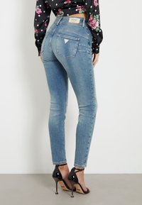 Žena v světle modrých skinny džínech s logem Guess na zadní kapse a černých vysokých podpatcích, stojící proti bílé zdi.