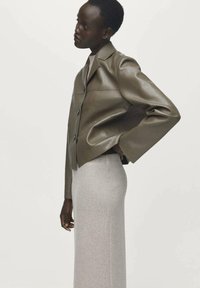 Femme portant une veste en cuir olive courte avec des boutons et une jupe gris clair taille haute, se tenant de profil sur un fond blanc.