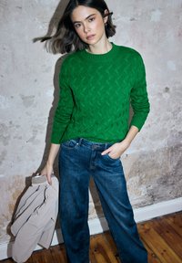 Groen gebreide trui met een gestructureerd patroon, gecombineerd met wijde, vervaagde blauwe jeans. Het model houdt een crème bomberjack in één hand vast.