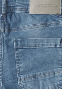 Jean en denim bleu clair avec une poche à rabat, présentant des détails de couture argentée et une étiquette beige indiquant "STREET ONE DENIM ATELIER."
