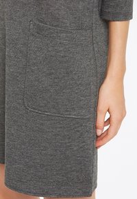 Robe grise avec une poche latérale, fabriquée dans un tissu doux au toucher tricoté. Des coutures apparentes accentuent l'ourlet et la poche.